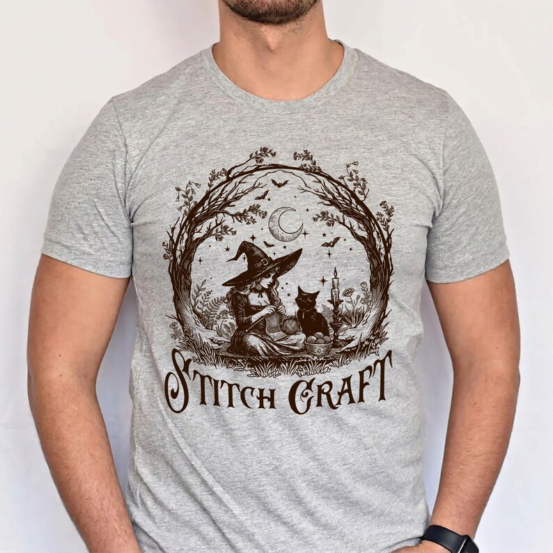 May include: A heather grey short-sleeve t-shirt featuring a dark brown graphic print of a witch knitting with a black cat. The design includes a crescent moon, stars, bats, and trees, with the text STITCH CRAFT printed in a stylised font below.