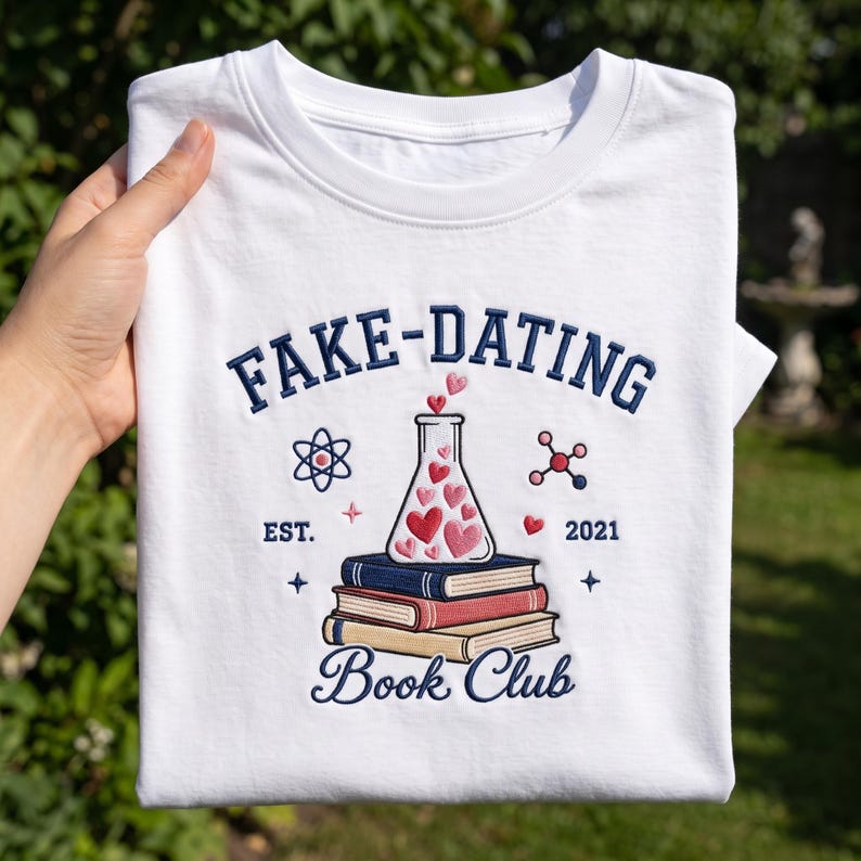 May include: A white t-shirt featuring an embroidered design with the text FAKE-DATING BOOK CLUB. The graphic includes a stack of three books, a laboratory flask filled with red and pink hearts, an atom symbol, and the text EST. 2021.