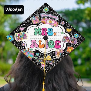 Op de afbeelding: Een afstudeerhoed met een zwarte achtergrond en witte spikkels, versierd met kleurrijke afbeeldingen. De hoed toont de tekst "MRS. ALICE" in grote, retro letters, omringd door bloemen, harten en andere decoratieve elementen. De hoed heeft een gouden kwast.