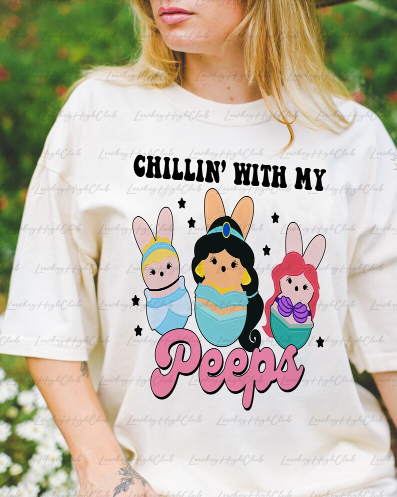 May include: Cream-colored t-shirt with the text "CHILLIN' WITH MY Peeps." The graphic features three cartoon characters in Easter egg shapes with bunny ears. The characters are in blue, teal, and pink, with black stars.