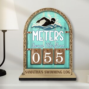 May include: A wooden swimming log with a teal background and a swimmer graphic. The log displays "METERS Swam This Year" and a counter showing "055". The base reads "SAMANTHA'S SWIMMING LOG".