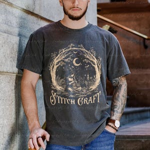 May include: A charcoal grey short-sleeve t-shirt featuring a beige graphic print of a witch sitting under a crescent moon and tree branches, with a cat and the text "STITCH CRAFT" centred below the illustration in a decorative serif font.