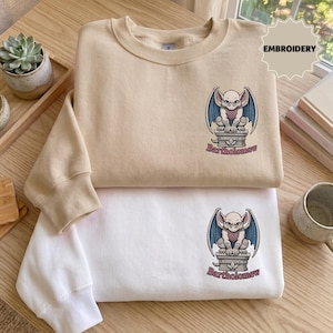 May include: A beige sweatshirt and a white sweatshirt, each featuring an embroidered graphic of a winged gargoyle perched on a stone pedestal with the text Bartholomew below it. The sweatshirts have long sleeves and a crew neckline design.
