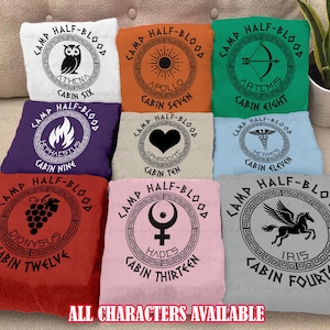 May include: A collection of colorful throw pillows, each featuring a circular design with text and a graphic. The pillows are in various colors, including white, orange, green, purple, tan, light blue, red, pink, and gray. The text reads "Camp Half-Blood" and cabin names.