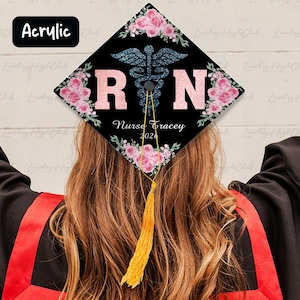 May include: A black graduation cap with pink floral accents and the letters "RN" in pink glitter. A blue caduceus symbol is centered above the letters. The text "Nurse Tracey 2024" is below the letters. A gold tassel hangs from the cap.