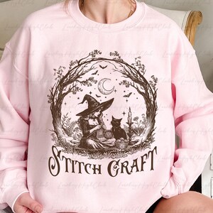May include: A light pink crewneck sweatshirt featuring a dark brown graphic print of a witch in a pointed hat knitting with a black cat nearby. The scene is framed by bare tree branches, a crescent moon, and bats, with the text STITCH CRAFT below.
