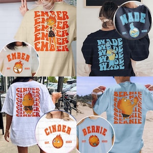 May include: Collection of t-shirts with various designs. One shirt features the name "EMBER" with a cartoon character. Another displays "WADE" with a cartoon. Others show "CINDER" and "BERNIE" with cartoon graphics. All shirts include "EST. 2023".