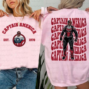 May include: A light pink sweatshirt featuring the words "CAPTAIN AMERICA" in red arched text. Below the text is a circular graphic with a portrait of a character. The back of the sweatshirt has a repeating pattern of the words "CAPTAIN AMERICA" and a full body image of the character.