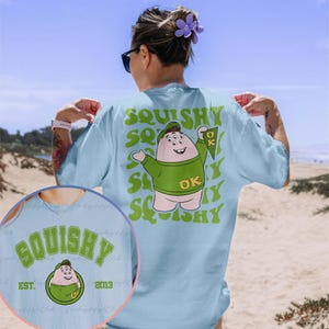 May include: Light blue t-shirt featuring a green cartoon character holding a purple pennant with the letters "OK" on it. The character has four eyes and is wearing a green shirt with the letters "OK" on it. The text "SQUISHY" is repeated in a wavy font on the shirt.