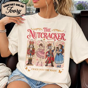 May include: An ivory t-shirt featuring a graphic print with the text The Nutcracker and Crack Into The Magic. The design includes four panels showing a nutcracker, a ballerina, a mouse king, and a girl holding a nutcracker doll.