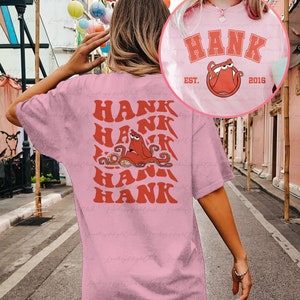 May include: Pink t-shirt with a red octopus graphic and the text "HANK" repeated multiple times. The octopus has a red body and white eyes. The text is in a retro font style. The shirt also has the text "HANK EST. 2016" on the front.