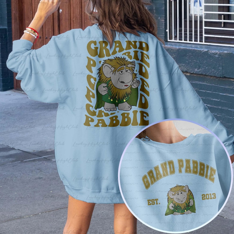 May include: Light blue sweatshirt with a graphic print of a cartoon character and the text "GRAND PABBIE". The character is green and brown and has a beard. The text is in a gold color.