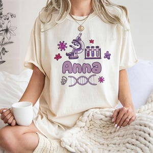 May include: Cream-colored t-shirt featuring a science-themed design. The design includes a microscope, test tubes, and the name "Anna" in purple. A DNA strand and other scientific elements are also present.