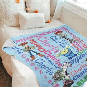 May include: A light blue fleece blanket featuring a grid pattern with the name Charlie repeated in red, pink, and blue text. The blanket displays cartoon characters Woody, Jessie, Buzz Lightyear, and an alien from the Toy Story film franchise.