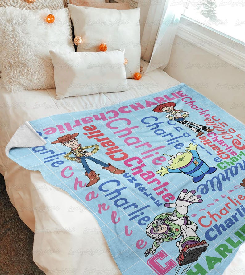 May include: A light blue fleece blanket featuring a grid pattern with the name Charlie repeated in red, pink, and blue text. The blanket displays cartoon characters Woody, Jessie, Buzz Lightyear, and an alien from the Toy Story film franchise.