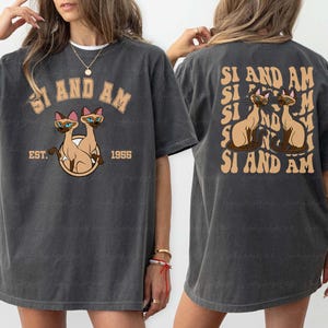 May include: Dark gray t-shirt featuring a Siamese cat graphic with the text "SI AND AM" and "EST. 1955". The back of the shirt repeats the graphic and text. The shirt is a casual, vintage style.
