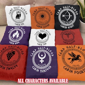 May include: A collection of colorful throw pillows with circular designs featuring Greek mythology symbols and text. Each pillow displays "Camp Half-Blood" and a cabin number, with symbols like an owl, sun, bow and arrow, and grapes. The text "ALL CHARACTERS AVAILABLE" is at the bottom.