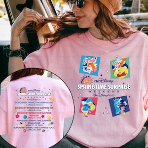 Könnte beinhalten: Hellrosa T-Shirt mit der Aufschrift "2026 runDisney Springtime Surprise Weekend Walt Disney World". Das Shirt hat Cartoon-Charakter-Grafiken. Auf der Rückseite des Shirts sind Rennveranstaltungen aufgeführt.