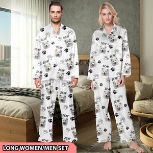 Könnte beinhalten: Passende weiße Pyjama-Sets mit einem sich wiederholenden Muster aus Hundegesicht, Pfotenabdrücken und dem Wort "BEAR". Das langärmlige Oberteil und die Hose werden von einem Mann und einer Frau getragen. Der Text "LONG WOMEN/MEN SET" befindet sich unten.