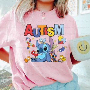 Op de afbeelding: Roze T-shirt met een blauw Stitch-personage uit Lilo & Stitch. Stitch is omringd door kleurrijke puzzelstukjes, vogels en een lint met een puzzelstukje. Het woord "Autisme" staat in regenboogkleuren boven Stitch.