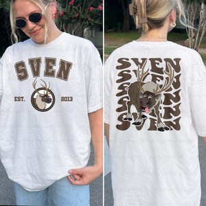 Pode incluir: Camiseta branca com um gr&aacute;fico marrom de uma rena chamada Sven, com o texto "SVEN" acima e "EST. 2013" abaixo. A parte de tr&aacute;s apresenta um gr&aacute;fico maior de Sven e o nome repetido em um padr&atilde;o ondulado. Uma camiseta unissex casual.