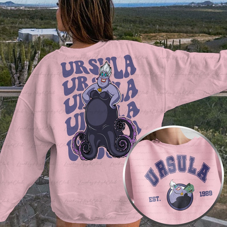 Pu&ograve; includere: Felpa rosa con una grafica di Ursula de La Sirenetta. La grafica presenta Ursula nella sua forma di polpo con il testo "Ursula" ripetuto in un font retr&ograve;.