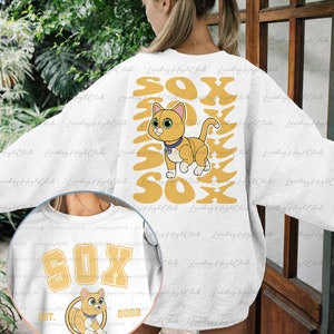 May include: White sweatshirt with a graphic of a cartoon orange cat with blue eyes and a yellow collar. The cat is surrounded by the word "SOX" in yellow letters. The sweatshirt also has a smaller graphic on the front with the word "SOX" in yellow letters and the text "EST. 2022" in black letters.
