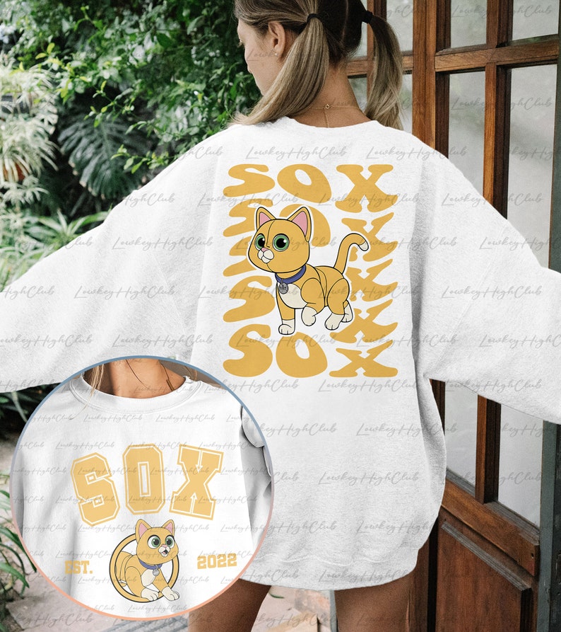 May include: White sweatshirt with a graphic of a cartoon orange cat with blue eyes and a yellow collar. The cat is surrounded by the word "SOX" in yellow letters. The sweatshirt also has a smaller graphic on the front with the word "SOX" in yellow letters and the text "EST. 2022" in black letters.