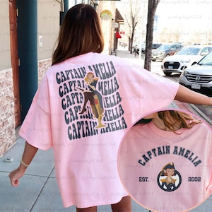 May include: Pink t-shirt featuring a graphic print of a woman wearing a captain's hat and the text "Captain Amelia" repeated in a retro font. The text "Captain Amelia" is also printed on the front of the shirt in a black arch with the text "Est. 2002" below.