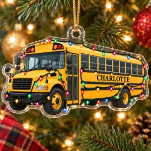 May include: A yellow school bus ornament featuring a string of colorful Christmas lights draped across the exterior. The name Charlotte is printed in black lettering on the side of the bus. The ornament is made of clear acrylic with a gold hanging cord.