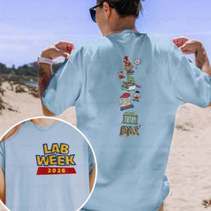 May include: Light blue t-shirt with the text "LAB WEEK 2026" in yellow and red. The back of the shirt features a vertical design of cartoon lab-related images in various colors, including a microscope, beakers, and a DNA strand.