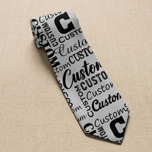 May include: A light gray necktie with the word "Custom" in various fonts and sizes, along with the letter "C" printed in black. The tie is angled, showcasing the design.