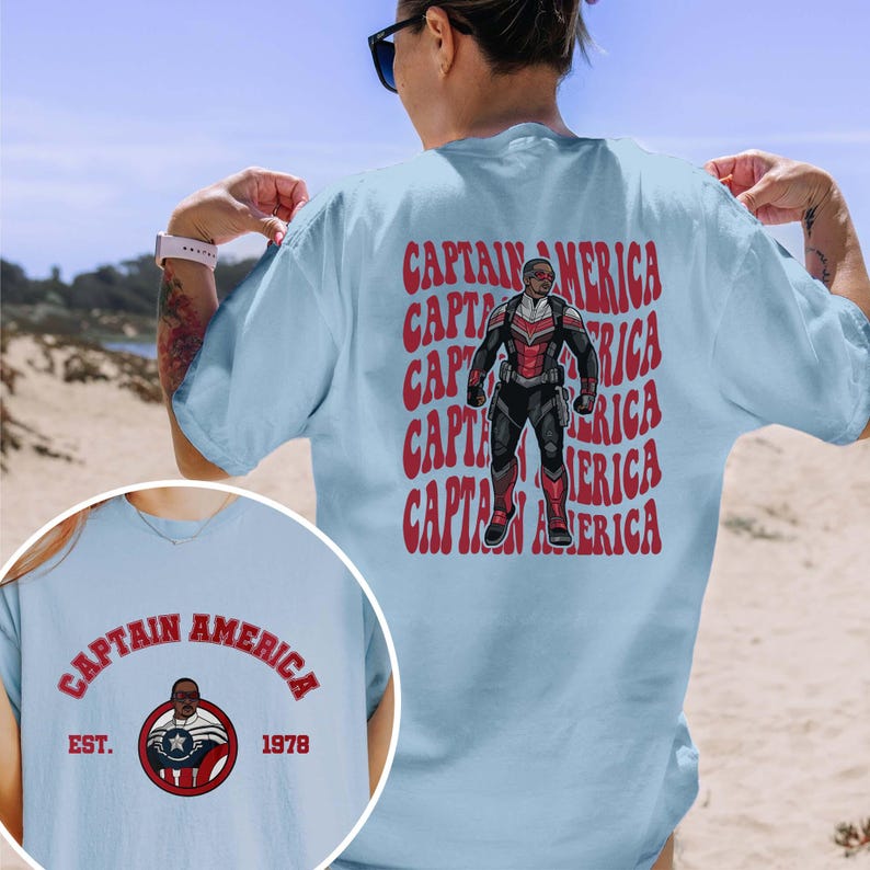 May include: Light blue t-shirt featuring a Captain America graphic on the back and a smaller circular design on the front. The back graphic includes the text "CAPTAIN AMERICA" repeated vertically with an illustration of a superhero. The front design includes the text "CAPTAIN AMERICA" and "EST. 1978".