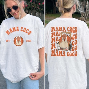 May include: White t-shirt featuring the text "MAMA COCO" in orange, with an illustration of an elderly woman in a wheelchair. The front of the shirt has the text "EST. 2021". The back of the shirt repeats "MAMA COCO" with the same illustration.