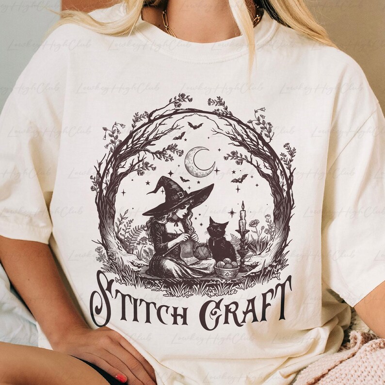 May include: A cream-coloured t-shirt featuring a dark brown graphic of a witch wearing a pointed hat, knitting with a black cat nearby. The scene is framed by tree branches, a crescent moon, and bats, with the text STITCH CRAFT printed below.