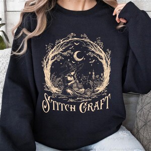 May include: A black crewneck sweatshirt featuring a beige graphic print of a witch in a pointed hat knitting with a black cat nearby. The design is framed by bare tree branches, a crescent moon, and bats, with the text STITCH CRAFT below.