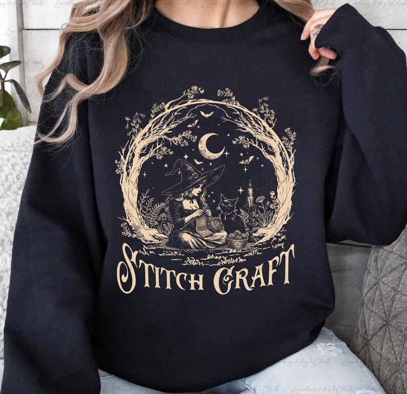 May include: A black crewneck sweatshirt featuring a beige graphic print of a witch in a pointed hat knitting with a black cat nearby. The design is framed by bare tree branches, a crescent moon, and bats, with the text STITCH CRAFT below.