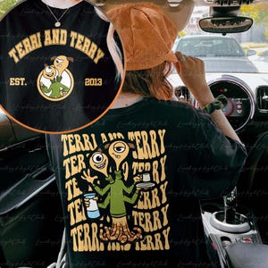 May include: Black t-shirt featuring a graphic print of two green and blue cartoon characters with the text "TERRI AND TERRY EST. 2013" on the front and the text "TERRI AND TERRY" repeated in a retro style on the back.