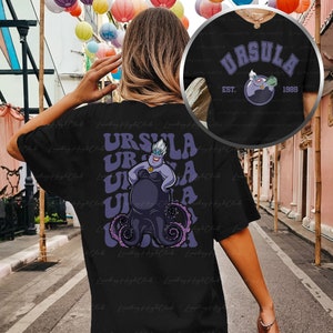 Pu&ograve; includere: Maglietta nera con una grafica viola e blu di Ursula de La Sirenetta. Il testo "URSULA EST. 1989" &egrave; stampato sul retro della maglietta.
