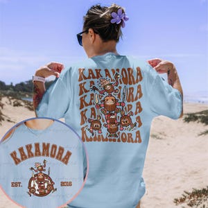 May include: A light blue t-shirt with a graphic print of a group of cartoon characters holding weapons. The text "Kakamora" is repeated in a wavy pattern on the shirt. The text "EST. 2016" is printed on the bottom left corner of the shirt.