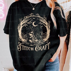 May include: A black short-sleeve t-shirt featuring a beige graphic of a witch wearing a pointed hat and knitting while sitting next to a black cat. The design includes a crescent moon, stars, and trees, with the text STITCH CRAFT printed below.