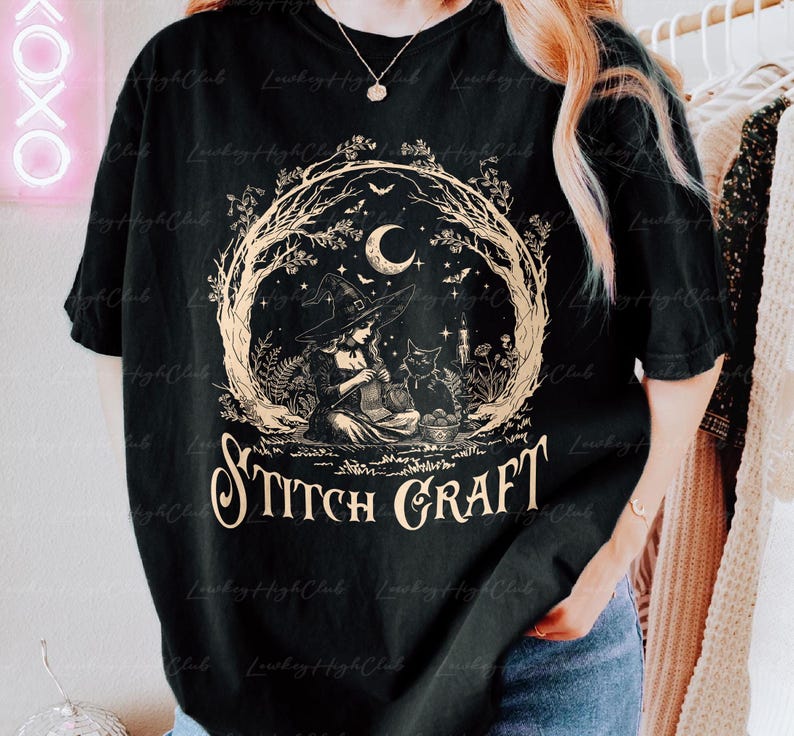 May include: A black short-sleeve t-shirt featuring a beige graphic of a witch wearing a pointed hat and knitting while sitting next to a black cat. The design includes a crescent moon, stars, and trees, with the text STITCH CRAFT printed below.