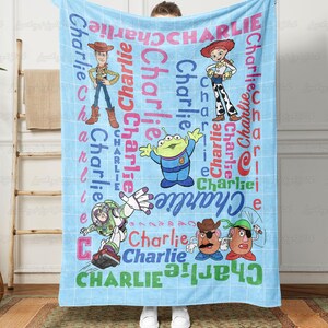 May include: A light blue fleece blanket featuring a white grid pattern and the name Charlie repeated in various colors and fonts. The design includes printed illustrations of Woody, Jessie, an alien, Buzz Lightyear, and Mr. Potato Head characters.