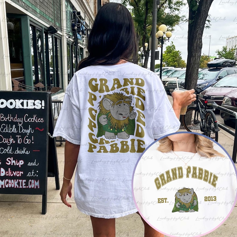 May include: White t-shirt with a green and gold graphic of a troll with the text "Grand Pabbie" on the back. The front of the shirt has the text "Grand Pabbie" in gold with the text "Est. 2013" underneath.