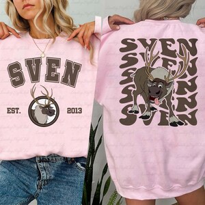 Pode incluir: Um moletom rosa claro com o nome "SVEN" em letras marrons e um gr&aacute;fico de rena. A frente do moletom tem o texto "EST. 2013". A parte de tr&aacute;s do moletom tem um grande gr&aacute;fico de rena e o nome "SVEN".
