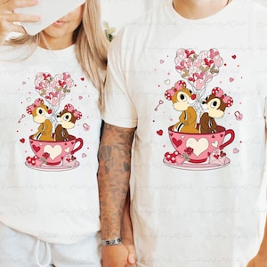 May include: White t-shirts featuring a cartoon illustration of two chipmunks in a pink teacup, holding heart-shaped balloons. The design includes hearts, keys, and an envelope, perfect for Valentine's Day or a romantic gift.