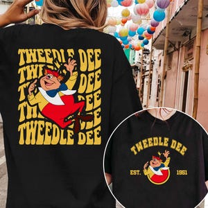 Könnte beinhalten: Schwarzes T-Shirt mit einer Cartoon-Illustration von Tweedle Dee aus Alice im Wunderland. Das Design zeigt die Figur in Rot, Gelb und Weiß, mit dem Text "TWEEDLE DEE" in Gelb. Ein kleineres Design ist ebenfalls sichtbar.