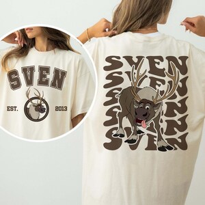 Pode incluir: Camiseta creme com um gr&aacute;fico marrom de Sven, a rena do filme Frozen. A frente da camiseta tem o nome "SVEN" acima de um emblema circular com Sven e o texto "EST. 2013". A parte de tr&aacute;s da camiseta repete "SVEN" em um padr&atilde;o ondulado.