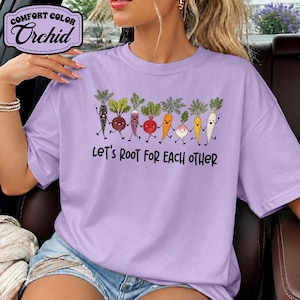 Può includere: Una t-shirt viola chiaro con una grafica di varie verdure colorate in stile cartone animato che si tengono per mano. Sotto le verdure, il testo "LET'S ROOT FOR EACH OTHER" è scritto in lettere nere. La t-shirt ha maniche corte.