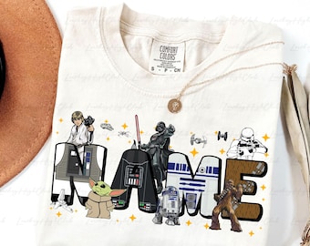 May include: Cream-colored t-shirt featuring a Star Wars-themed design. The word "NAME" is spelled out with Star Wars characters and imagery. Includes characters like Darth Vader, R2-D2, and Chewbacca. A gold necklace and a brown hat are visible.